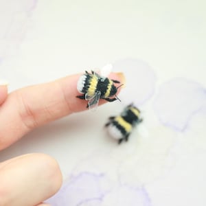 May include: Two miniature bumblebees, featuring black and yellow striped bodies and translucent wings. One bee rests on a fingertip, while the other is nearby. The background is a soft, watercolor-style wash of purple and white.