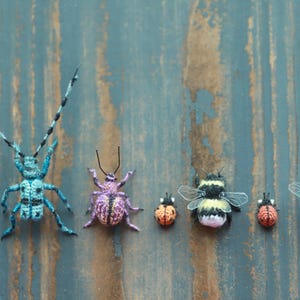 May include: A selection of handcrafted crochet insects, featuring a large blue beetle, a purple spider, two ladybirds, and two bumblebees. The insects are displayed on a weathered blue wooden surface.