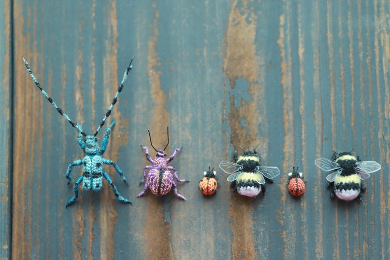 May include: A selection of handcrafted crochet insects, featuring a large blue beetle, a purple spider, two ladybirds, and two bumblebees. The insects are displayed on a weathered blue wooden surface.