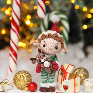 May include: A crocheted elf doll wearing a red and white hat, green and white striped outfit, and red mittens, holding a small reindeer. Candy canes and Christmas ornaments are in the background. The doll is approximately 4 inches tall.