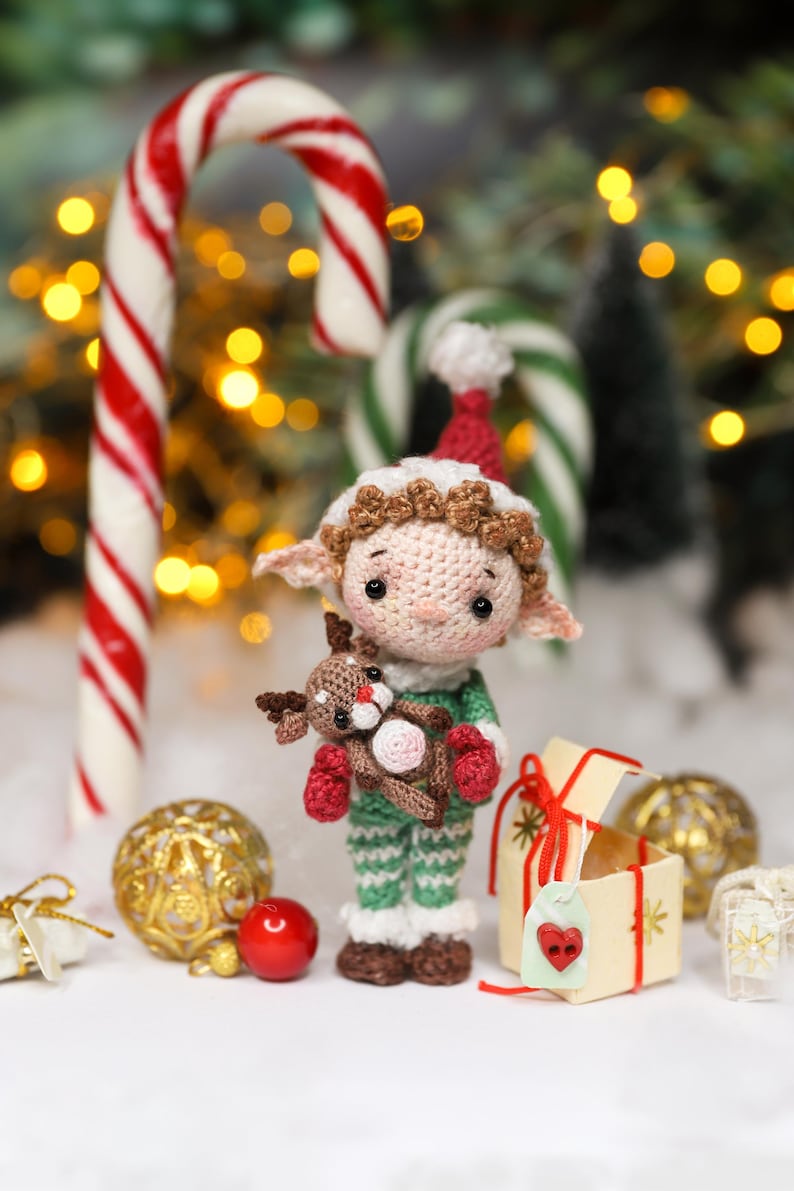 May include: A crocheted elf doll wearing a red and white hat, green and white striped outfit, and red mittens, holding a small reindeer. Candy canes and Christmas ornaments are in the background. The doll is approximately 4 inches tall.