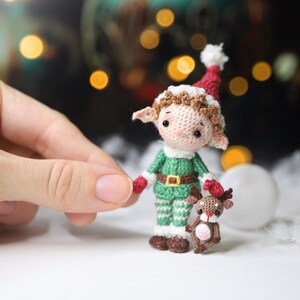 May include: A crocheted elf doll wearing a green outfit with a red and white hat, holding a small crocheted reindeer. The elf has brown hair and a gold belt. The reindeer is brown with a pink belly. Christmas theme.