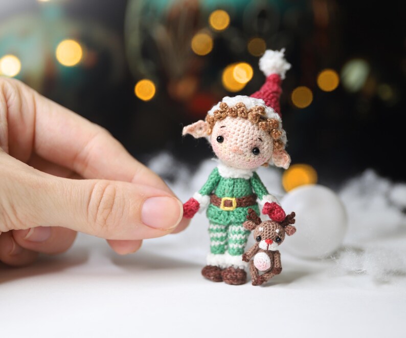 May include: A crocheted elf doll wearing a green outfit with a red and white hat, holding a small crocheted reindeer. The elf has brown hair and a gold belt. The reindeer is brown with a pink belly. Christmas theme.