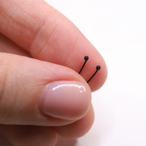 May include: Close-up of two small, black, straight pins with rounded heads. The pins are held between two fingers against a white background. The pins are likely used for crafting or sewing projects.
