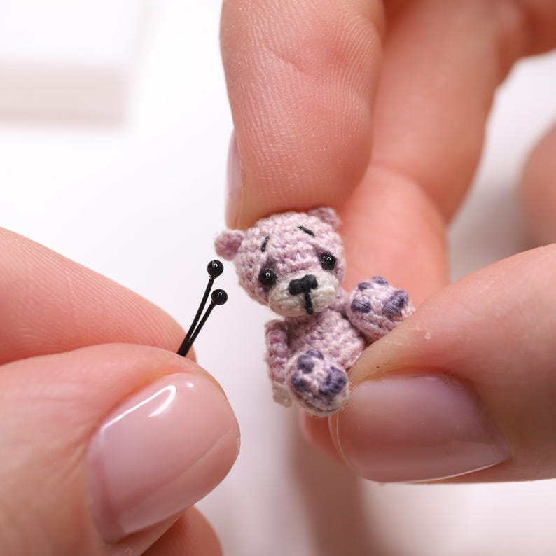 May include: A miniature, hand-crocheted teddy bear in shades of lavender and purple. The tiny bear has a black nose and eyes, and is being held between two fingers. Two black pins are in the foreground.