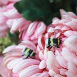 May include: Close-up of a pink flower arrangement with two small, decorative bees. The flower petals are a soft pink, and the bees have black and yellow striped bodies. Green leaves are visible in the background.