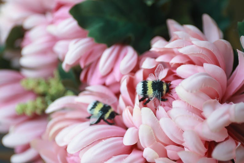 May include: Close-up of a pink flower arrangement with two small, decorative bees. The flower petals are a soft pink, and the bees have black and yellow striped bodies. Green leaves are visible in the background.