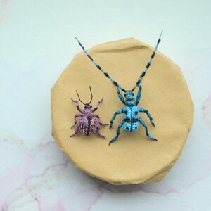 May include: Two handmade crochet insects rest on a tan, irregularly shaped surface. One is a small, purple beetle with black antennae. The other is a larger, blue insect with long antennae and a black and blue pattern.