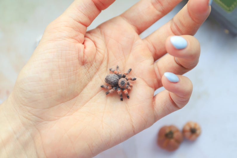 May include: A miniature, detailed spider figurine with a brown and orange body and legs, resting in an open palm. The spider's body is a mix of brown and gray, with orange markings on its legs. The hand has light blue nail polish.
