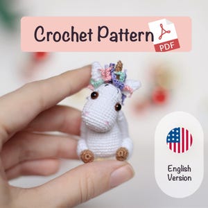 May include: A small, white crocheted unicorn with a colorful mane and a golden horn. The unicorn is held in a hand, with the text "Crochet Pattern PDF" in a pink banner above. An "English Version" label is also visible.
