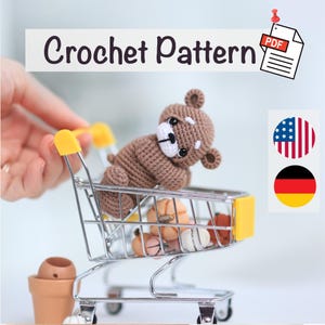 Könnte beinhalten: Ein gehäkelter brauner Teddybär sitzt in einem Miniatur-Einkaufswagen mit gelben Akzenten. Der Wagen enthält auch kleine, runde, hellfarbige Objekte. Die Worte "Crochet Pattern" werden über dem Wagen zusammen mit einem PDF-Symbol angezeigt.