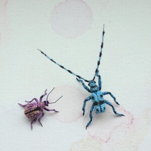May include: Two handmade insect sculptures. One is a small, purple and gold beetle. The other is a larger, blue and black longhorn beetle with long antennae. Both are set against a white background with pink watercolour-like accents.