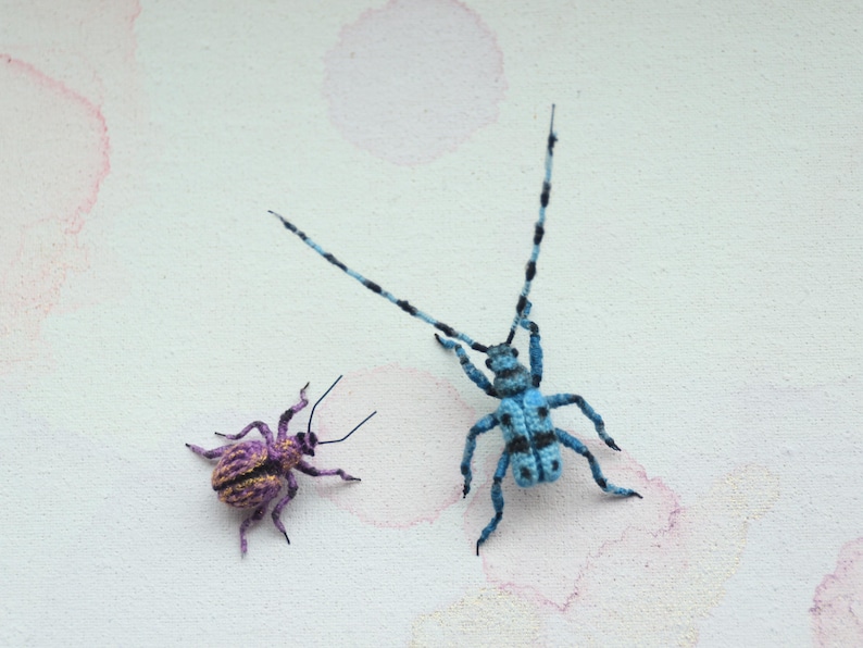 May include: Two handmade insect sculptures. One is a small, purple and gold beetle. The other is a larger, blue and black longhorn beetle with long antennae. Both are set against a white background with pink watercolour-like accents.
