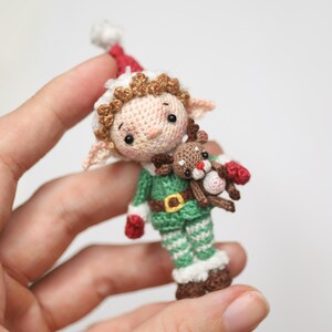 May include: A handmade crochet elf doll wearing a green and white outfit with a red hat and mittens. The elf is holding a small reindeer. The doll has brown hair and a belt with a gold buckle. A festive holiday decoration.