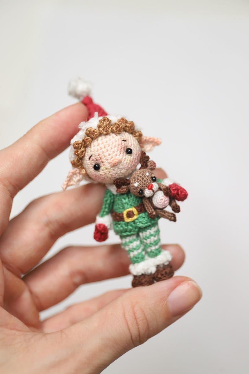 May include: A handmade crochet elf doll wearing a green and white outfit with a red hat and mittens. The elf is holding a small reindeer. The doll has brown hair and a belt with a gold buckle. A festive holiday decoration.