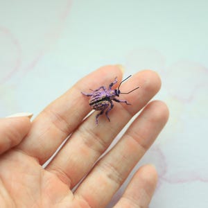 May include: A small, purple and black insect model with long antennae is placed on a finger. The insect features a striped abdomen and detailed legs. The background is a soft, light pink and white. The insect is approximately 1 cm in length.