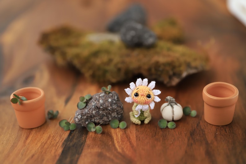 Puede incluir: Una escena caprichosa con una figura de girasol de ganchillo, peque&ntilde;as macetas de terracota, una roca y una calabaza en miniatura. El girasol tiene un centro amarillo y p&eacute;talos blancos. Plantas verdes y peque&ntilde;as esferas verdes est&aacute;n dispersas.