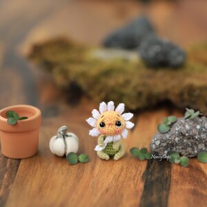 Puede incluir: Una mu&ntilde;eca de margarita de ganchillo hecha a mano con una cara sonriente, vestida con un atuendo verde, se sienta junto a una peque&ntilde;a calabaza, una maceta de terracota con una planta y una roca. La mu&ntilde;eca tiene p&eacute;talos blancos y un centro amarillo.