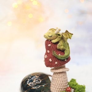 Miniature sleeping crochet dragon perched on crocheted red mushroom ring, tiny green amigurumi fantasy creature with folded wings and curled tail, detailed micro crochet toy shown on finger for scale.