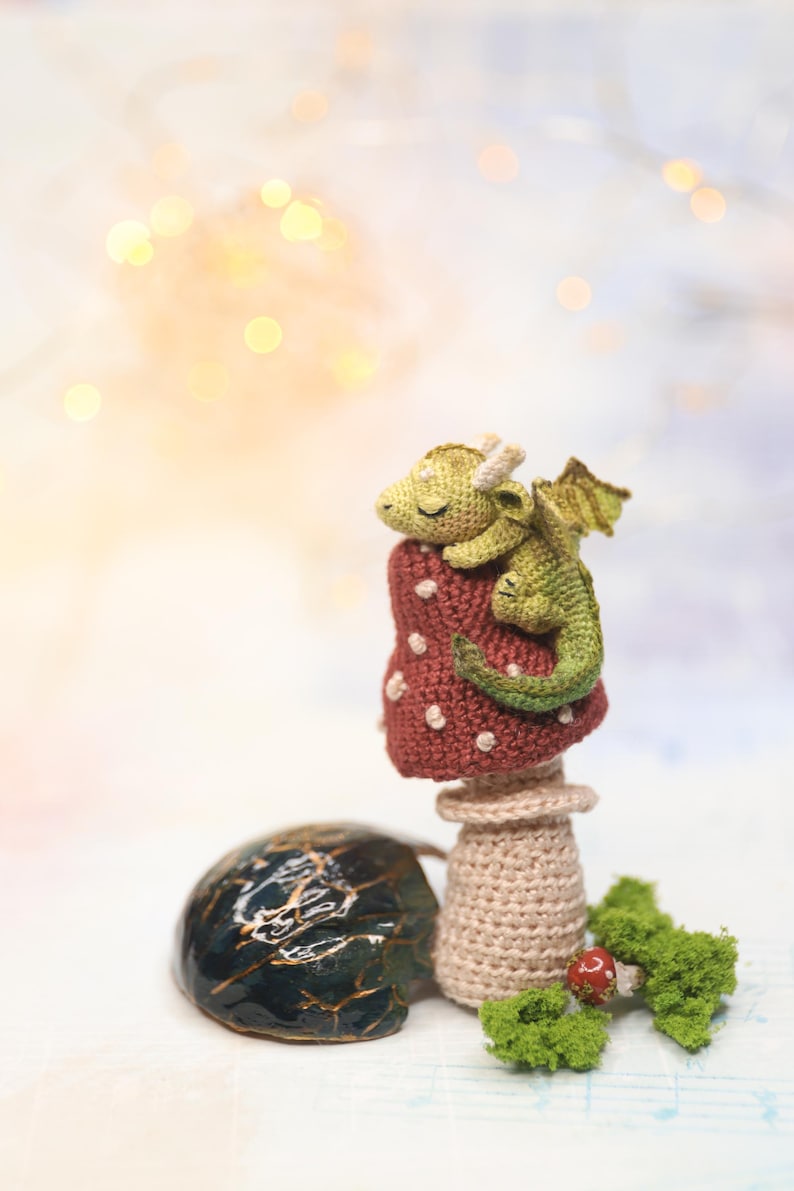 Miniature sleeping crochet dragon perched on crocheted red mushroom ring, tiny green amigurumi fantasy creature with folded wings and curled tail, detailed micro crochet toy shown on finger for scale.