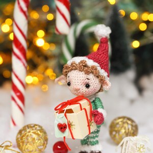 May include: A crocheted elf doll wearing a red hat and green outfit, holding a small gift box tied with red ribbon. A large candy cane with red and white stripes stands behind the doll, with Christmas ornaments and a small gift box in the foreground.