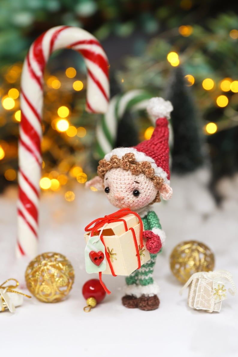 May include: A crocheted elf doll wearing a red hat and green outfit, holding a small gift box tied with red ribbon. A large candy cane with red and white stripes stands behind the doll, with Christmas ornaments and a small gift box in the foreground.