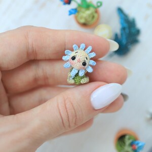 Puede incluir: Una figura de girasol en miniatura tejida a ganchillo con una cara sonriente, p&eacute;talos azules y una base verde. La artesan&iacute;a hecha a mano se sostiene en una mano, con otras plantas en miniatura en el fondo. La figura mide aproximadamente 2,5 cm de altura.