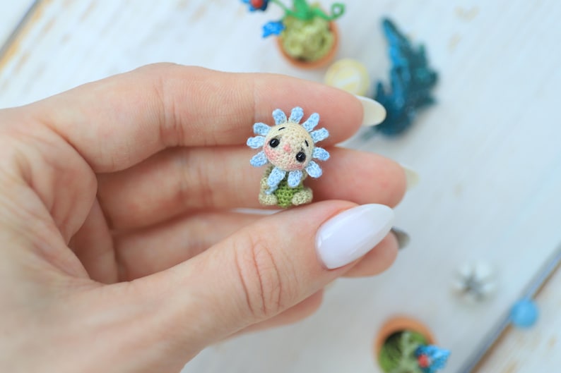 Puede incluir: Una figura de girasol en miniatura tejida a ganchillo con una cara sonriente, p&eacute;talos azules y una base verde. La artesan&iacute;a hecha a mano se sostiene en una mano, con otras plantas en miniatura en el fondo. La figura mide aproximadamente 2,5 cm de altura.