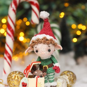 May include: A crocheted Christmas elf doll wearing a red hat, green outfit, and holding a gift box with a reindeer inside. The scene includes candy canes, gold ornaments, and a festive backdrop.