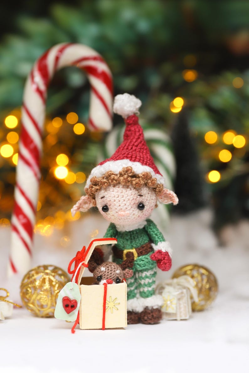 May include: A crocheted Christmas elf doll wearing a red hat, green outfit, and holding a gift box with a reindeer inside. The scene includes candy canes, gold ornaments, and a festive backdrop.