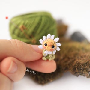 Puede incluir: Una peque&ntilde;a mu&ntilde;eca de margarita de ganchillo hecha a mano con una cara sonriente, p&eacute;talos blancos y un tallo verde. La mu&ntilde;eca se sostiene entre los dedos, con una madeja de hilo verde en el fondo. La cara de la mu&ntilde;eca tiene mejillas rosadas y ojos negros.