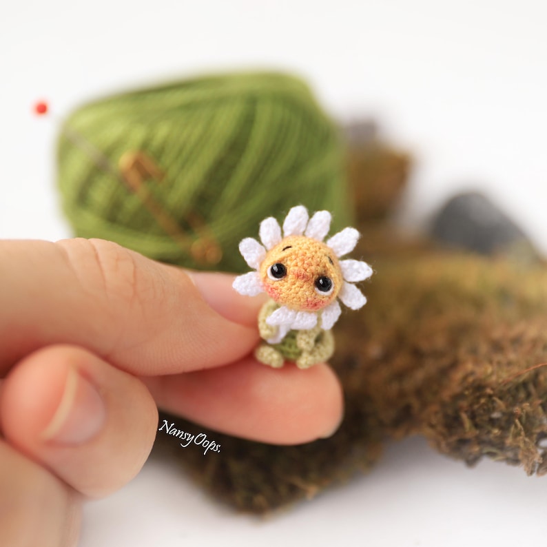 Puede incluir: Una peque&ntilde;a mu&ntilde;eca de margarita de ganchillo hecha a mano con una cara sonriente, p&eacute;talos blancos y un tallo verde. La mu&ntilde;eca se sostiene entre los dedos, con una madeja de hilo verde en el fondo. La cara de la mu&ntilde;eca tiene mejillas rosadas y ojos negros.