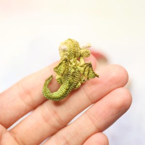 Tiny green micro crochet dragon resting on fingers, handmade amigurumi miniature with curled tail, small wings and horns, sleeping fantasy creature made with fine thread, scale shown by human hand.