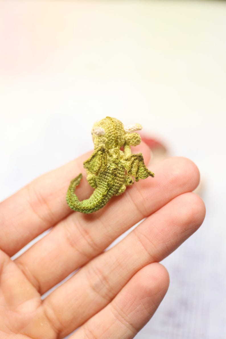 Tiny green micro crochet dragon resting on fingers, handmade amigurumi miniature with curled tail, small wings and horns, sleeping fantasy creature made with fine thread, scale shown by human hand.