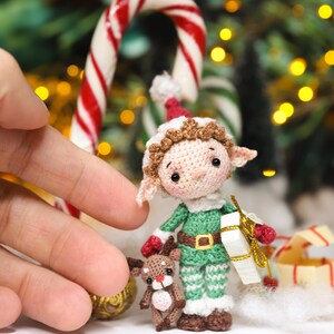May include: A handmade crochet elf doll with a green and white outfit, holding a small gift box. The elf is accompanied by a tiny reindeer. A red and white candy cane is in the background, suggesting a Christmas theme.