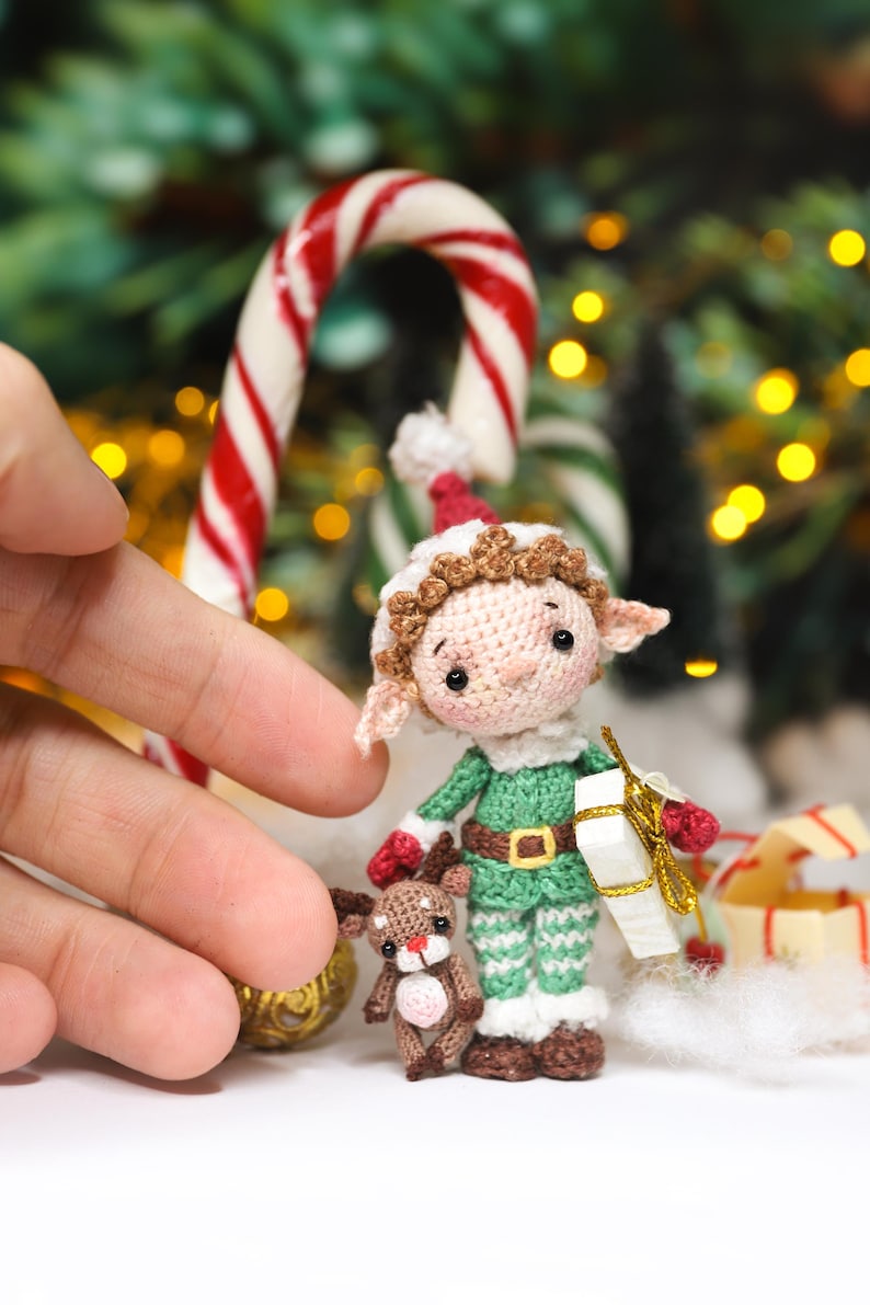 May include: A handmade crochet elf doll with a green and white outfit, holding a small gift box. The elf is accompanied by a tiny reindeer. A red and white candy cane is in the background, suggesting a Christmas theme.