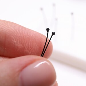 May include: Close-up of two black head pins held between fingers. The pins have a thin, straight wire with a small, round, black head. Other pins are visible in the background, out of focus, against a white backdrop.