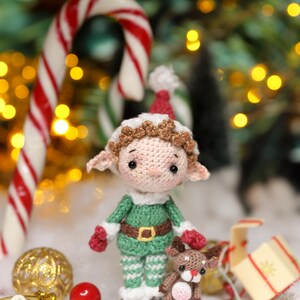May include: A crocheted Christmas elf doll wearing a green and white outfit with a red hat and mittens. The elf is holding a small crocheted reindeer. Candy canes and Christmas ornaments are in the background.