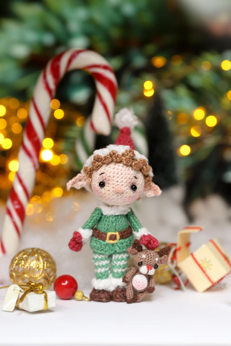 May include: A crocheted Christmas elf doll wearing a green and white outfit with a red hat and mittens. The elf is holding a small crocheted reindeer. Candy canes and Christmas ornaments are in the background.