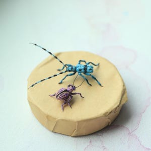 May include: Two crocheted insect figures are displayed on a round, tan cardboard base. A blue insect with long antennae and a purple insect are shown. The insects have black detailing, and the base sits on a light background.