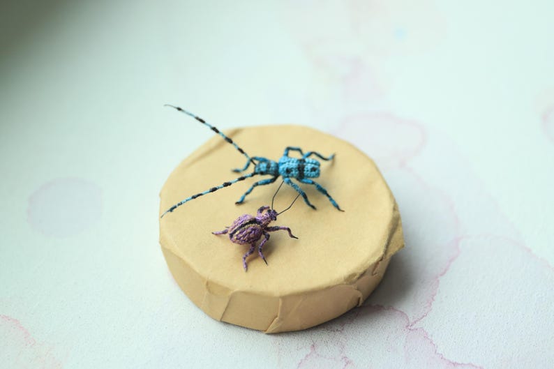 May include: Two crocheted insect figures are displayed on a round, tan cardboard base. A blue insect with long antennae and a purple insect are shown. The insects have black detailing, and the base sits on a light background.