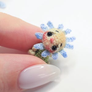 Puede incluir: Un girasol de ganchillo en miniatura con una cara sonriente. La flor tiene un centro amarillo, p&eacute;talos azules y una base verde. La cara presenta ojos negros y un rubor rosado. La artesan&iacute;a se sostiene entre los dedos.