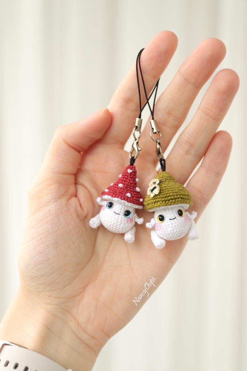 Pu&ograve; includere: Due ciondoli a forma di fungo all'uncinetto con facce sorridenti e cordini neri. Un fungo ha un cappello rosso a pois bianchi, l'altro un cappello verde. I ciondoli sono bianchi con guance rosa e piccole braccia e gambe.