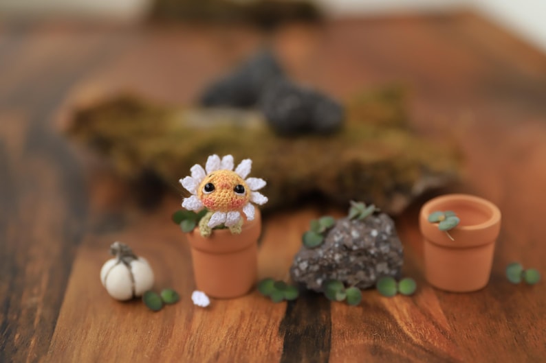 Puede incluir: Una criatura de ganchillo con forma de margarita se encuentra en una peque&ntilde;a maceta de terracota, rodeada de elementos de jard&iacute;n en miniatura. Tambi&eacute;n hay una peque&ntilde;a calabaza blanca y una roca con vegetaci&oacute;n. La escena est&aacute; ambientada sobre una superficie de madera.