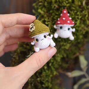 Pu&ograve; includere: Due piccole figure di funghi all'uncinetto. Un fungo ha un corpo bianco e un cappello verde con un fiore. L'altro fungo ha un corpo bianco e un cappello rosso con pois bianchi. Entrambi hanno facce sorridenti e piccole braccia e gambe.