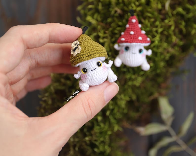 Pu&ograve; includere: Due piccole figure di funghi all'uncinetto. Un fungo ha un corpo bianco e un cappello verde con un fiore. L'altro fungo ha un corpo bianco e un cappello rosso con pois bianchi. Entrambi hanno facce sorridenti e piccole braccia e gambe.