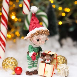 May include: A crocheted Christmas elf doll wearing a red and white hat and green outfit, holding a small gift box with a reindeer inside. The scene includes candy canes, ornaments, and a festive backdrop.