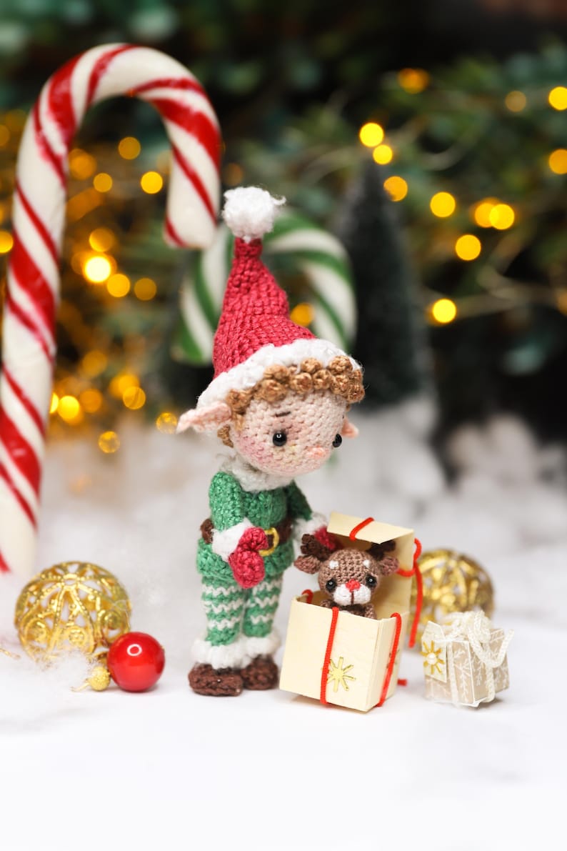 May include: A crocheted Christmas elf doll wearing a red and white hat and green outfit, holding a small gift box with a reindeer inside. The scene includes candy canes, ornaments, and a festive backdrop.