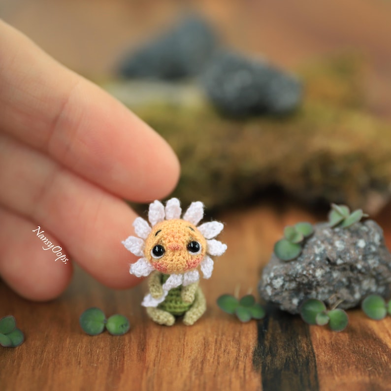 Puede incluir: Una mu&ntilde;eca de girasol de ganchillo hecha a mano con una cara sonriente, p&eacute;talos blancos y un cuerpo verde. La mu&ntilde;eca es peque&ntilde;a, con un dise&ntilde;o detallado, y est&aacute; colocada sobre una superficie de madera con peque&ntilde;as rocas y vegetaci&oacute;n. El nombre del artista, "NansyOops", es visible.