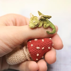 Miniature sleeping crochet dragon perched on crocheted red mushroom ring, tiny green amigurumi fantasy creature with folded wings and curled tail, detailed micro crochet toy shown on finger for scale.